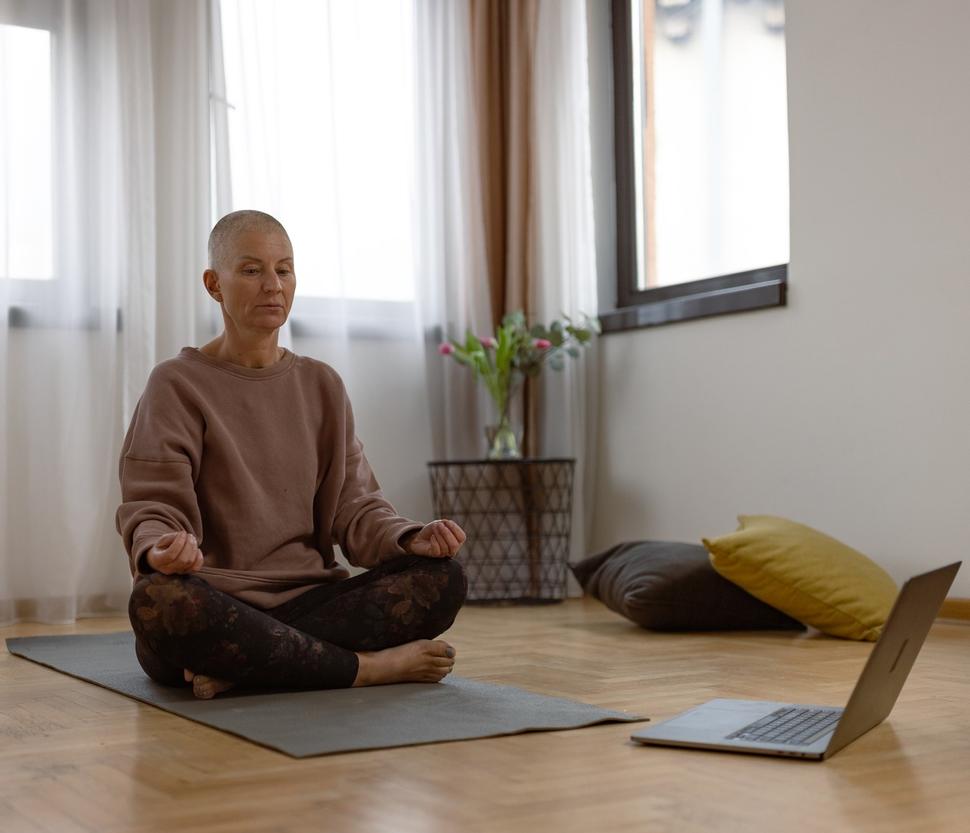 Mujer calva de mediana edad, sentada con las piernas cruzadas sobre una alfombra de yoga frente a una computadora portátil.