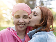 Photo of a smiling woman wearing a pink scarf on her head. Her eyes are closed and she is being embraced and kissed on the cheek by a woman with red hair.