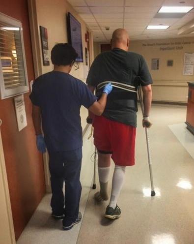 Photo taken from behind Ken Scott and his physical therapist in a hospital hallway. The physical therapist has his hand on Ken's back and Ken is using crutches to help him walk.