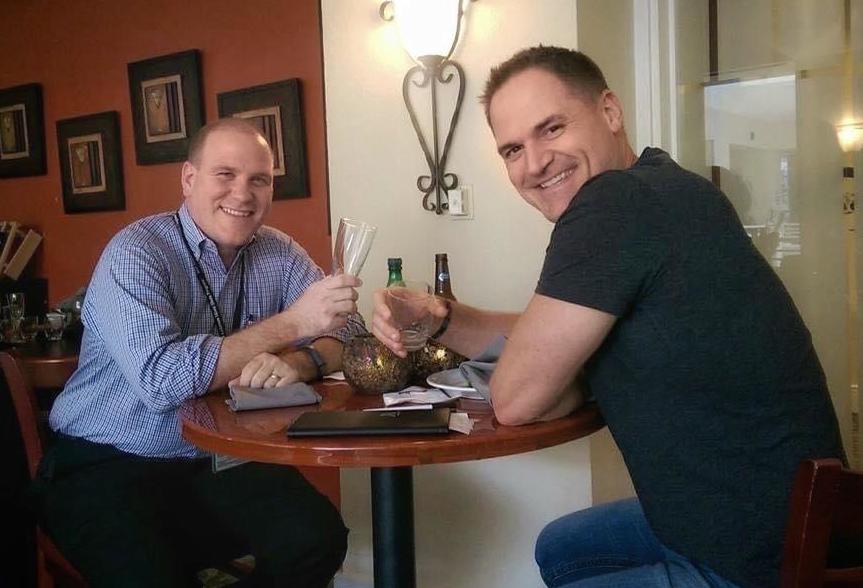 Tim and Ken sitting at a table, smiling with drinks in their hands