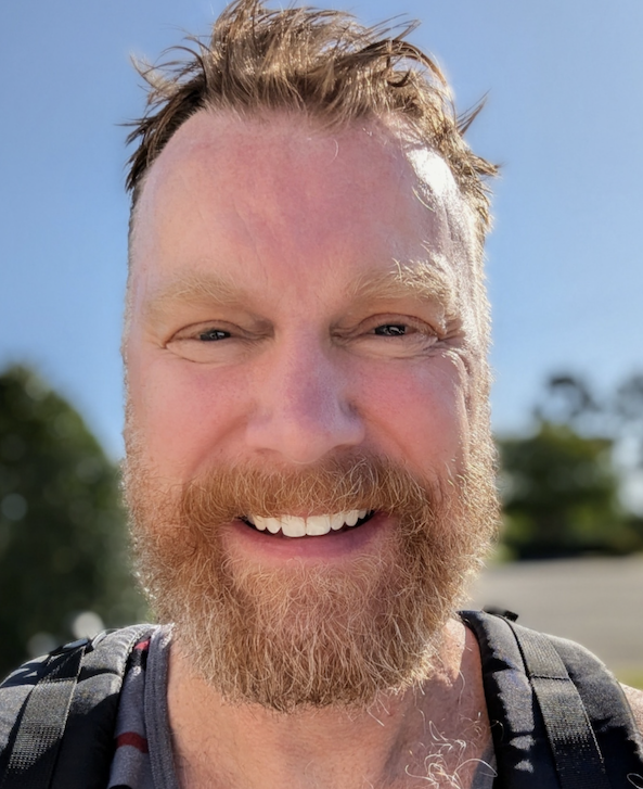 Hombre sonriente con barba tomándose una selfie al aire libre, con una mochila.