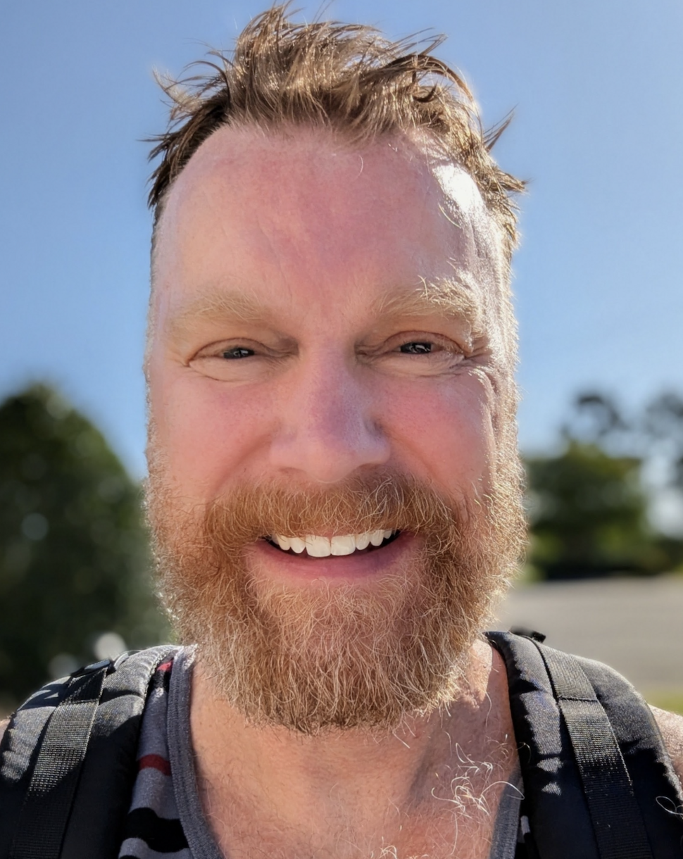 Smiling man with a beard taking an outdoor selfie, wearing a backpack over a striped tank top.