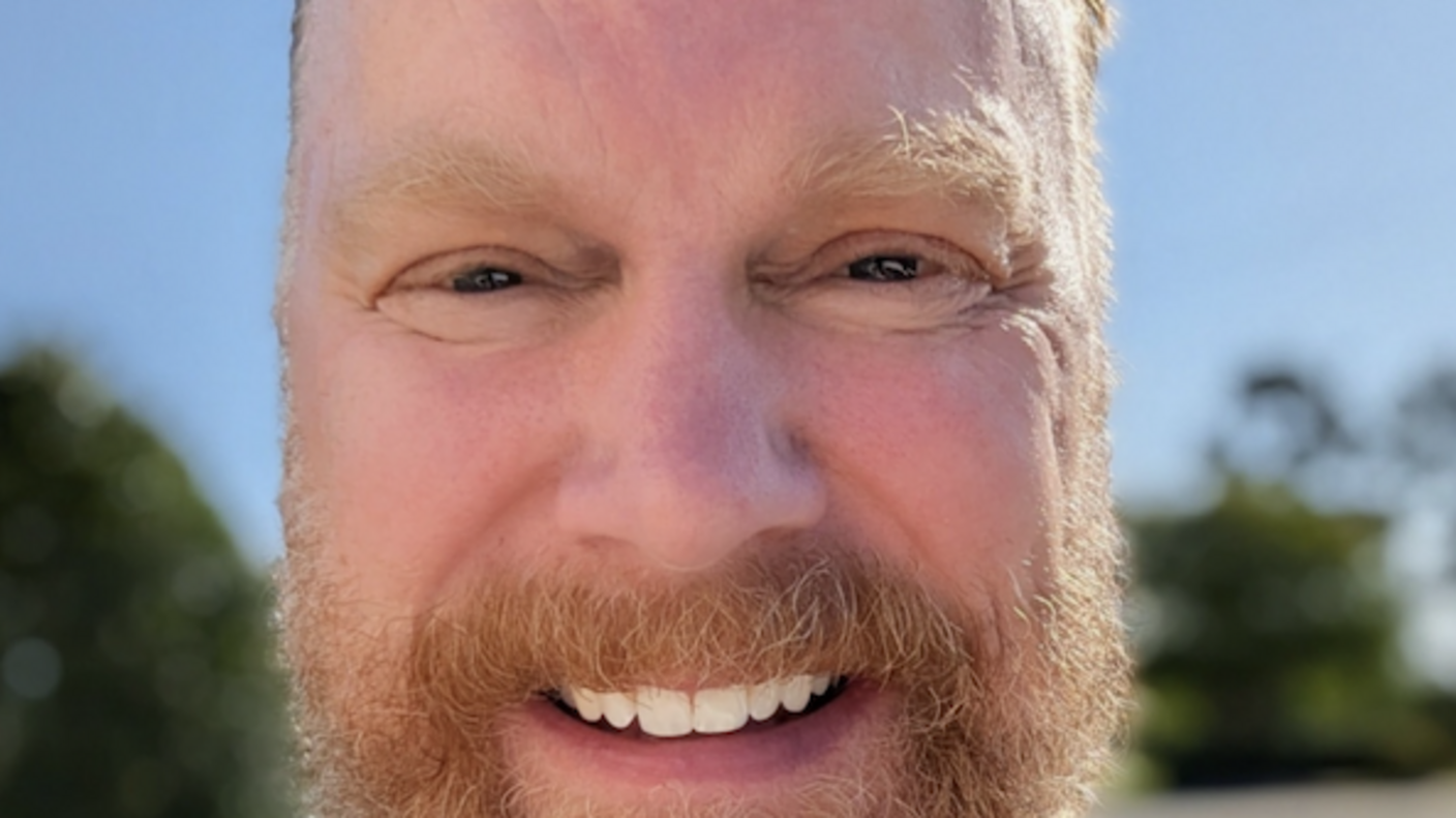 Smiling man with a beard taking an outdoor selfie, wearing a backpack over a striped tank top.