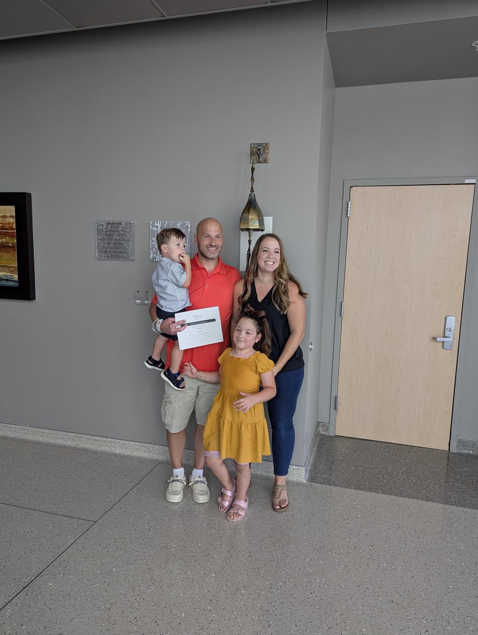 Smiling family of four with mom, dad (Josh), and their two young children standing beside the chemotherapy bell at a cancer treatment facility, celebrating dad finishing treatment, happy faces, hopeful moment.