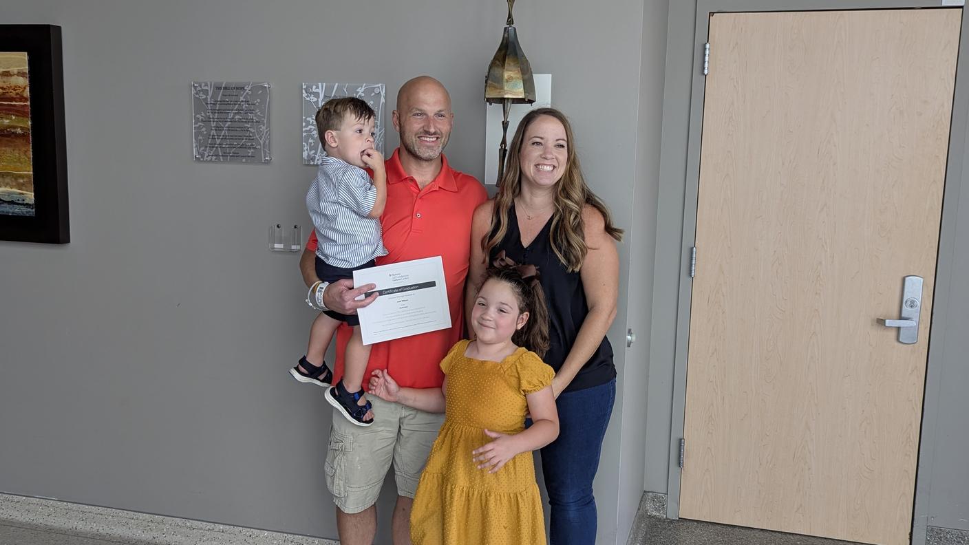 Smiling family of four with mom, dad (Josh), and their two young children standing beside the chemotherapy bell at a cancer treatment facility, celebrating dad finishing treatment, happy faces, hopeful moment.