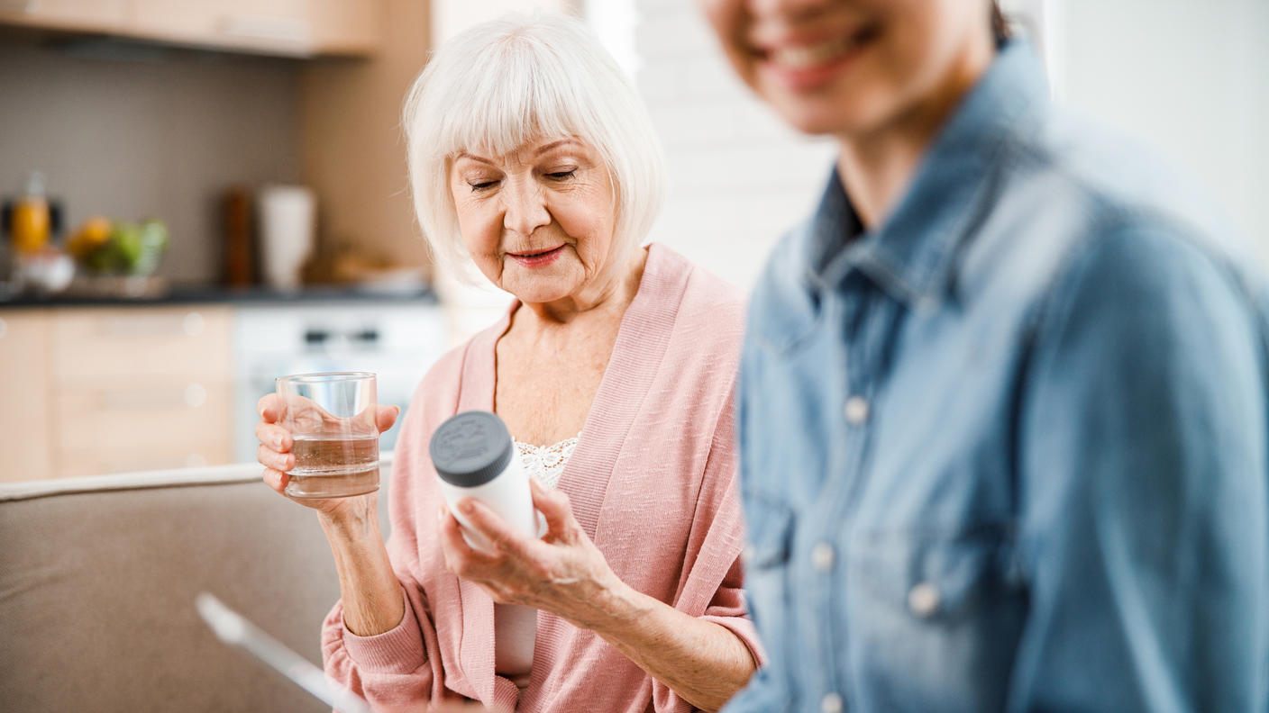 Older woman holding and reading pill bottle with a glass of water and younger woman looking at her phone.