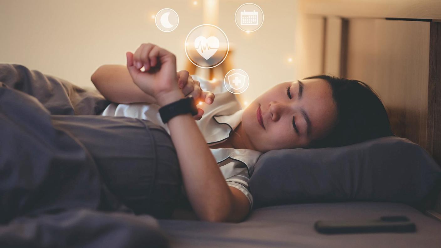 Woman lying in bed looking at her watch, which displays moon, heartbeat, calendar, and medical icons