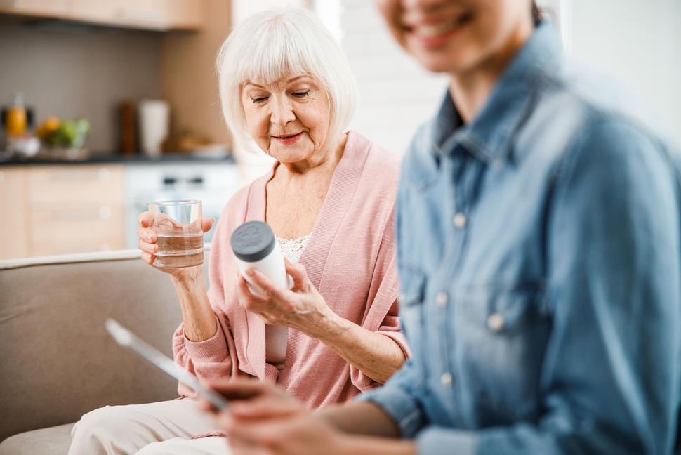 Older woman holding and reading pill bottle with a glass of water and younger woman looking at her phone.