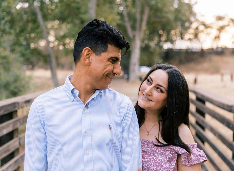 Liz and Julio smiling at one another while standing on a bridge during sunset