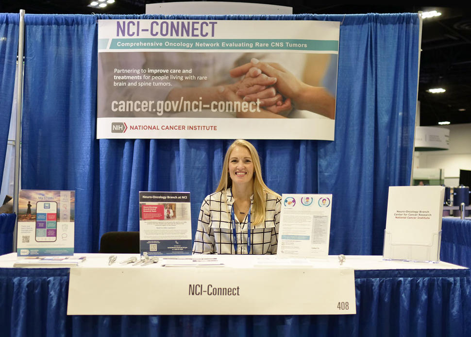 Woman smiling at a conference booth with an "NCI-CONNECT" sign and informational pamphlets on a table