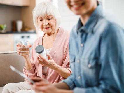 Older woman holding and reading pill bottle with a glass of water and younger woman looking at her phone.