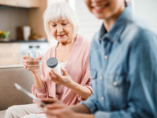 Older woman holding and reading pill bottle with a glass of water and younger woman looking at her phone.