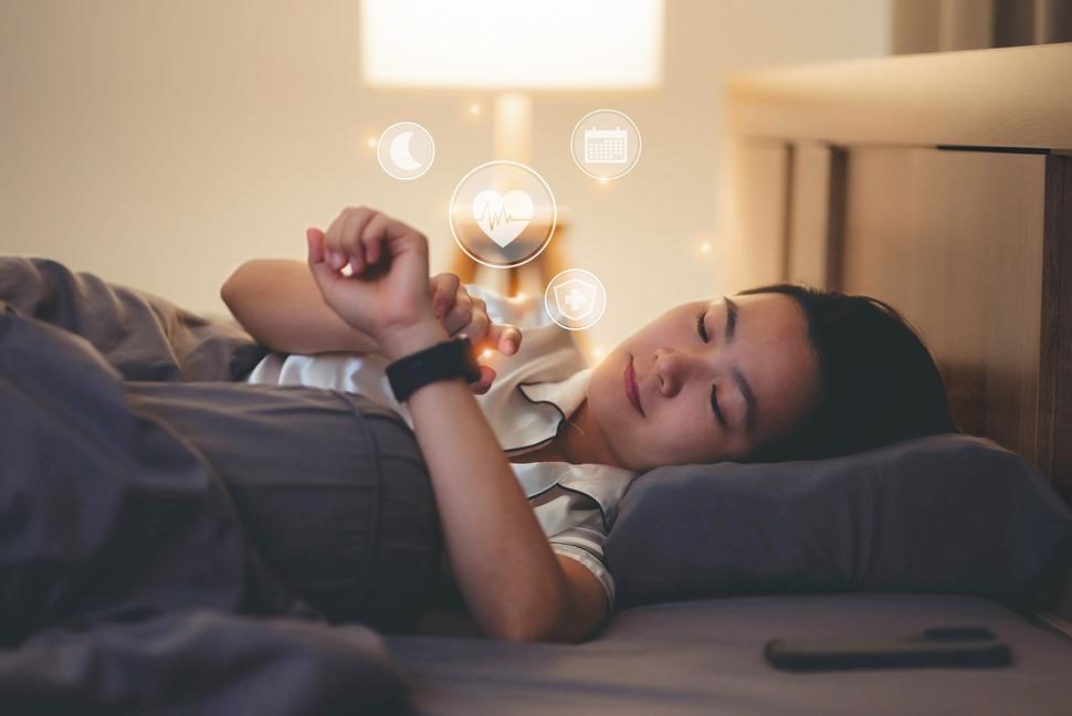 Woman lying in bed looking at her watch, which displays moon, heartbeat, calendar, and medical icons