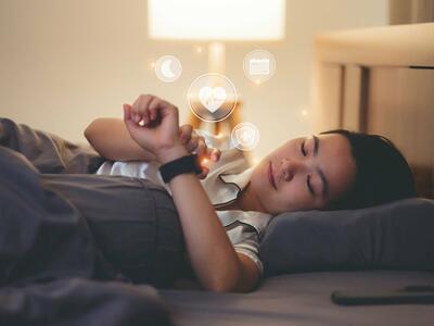 Woman lying in bed looking at her watch, which displays moon, heartbeat, calendar, and medical icons