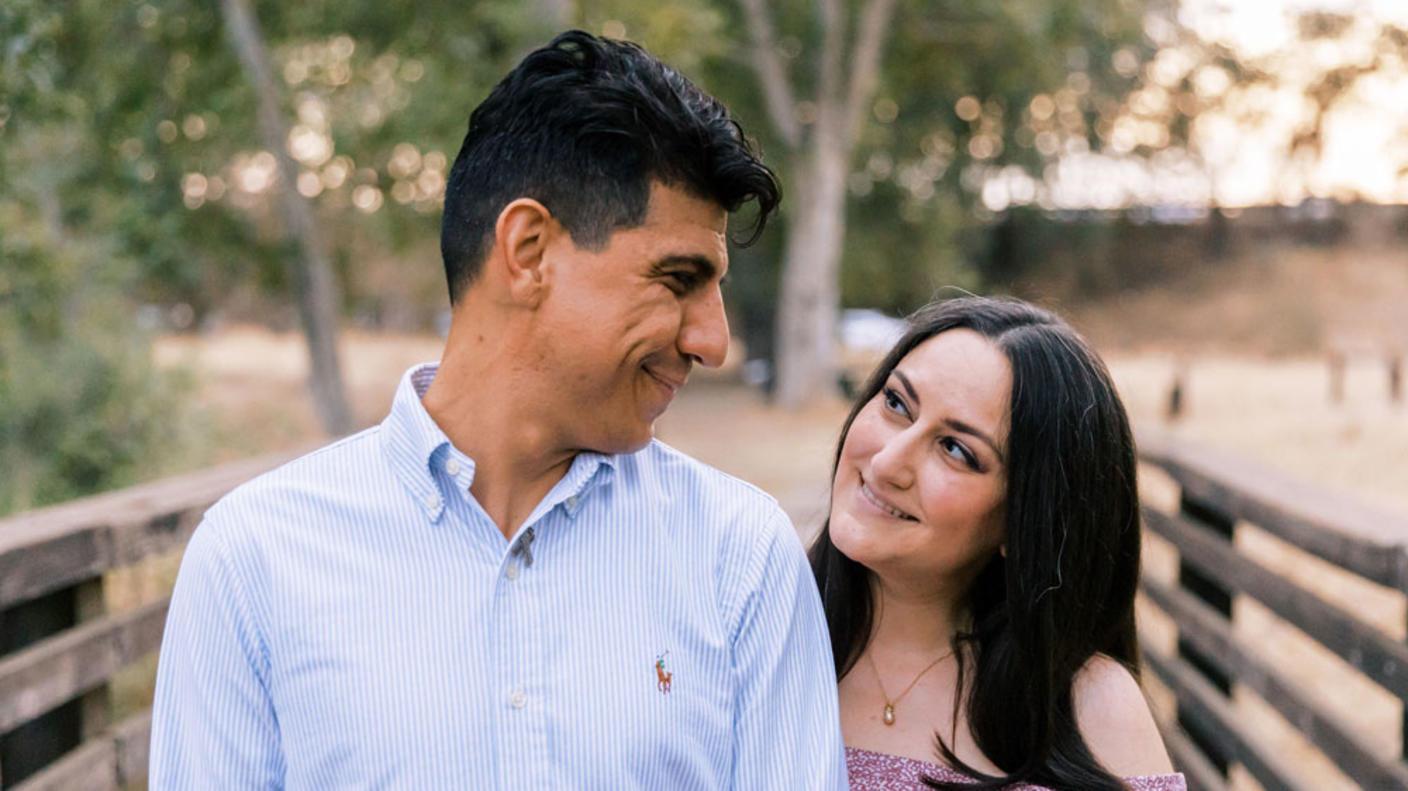Liz and Julio smiling at one another while standing on a bridge during sunset