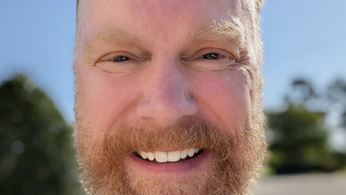 Smiling man with a beard taking an outdoor selfie, wearing a backpack over a striped tank top.