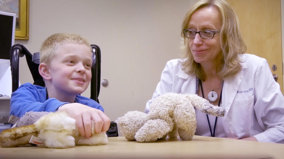 Brigitte Widemann (a light-skinned, blonde, female doctor in a white lab coat) is smiling and looking at a boy in a blue, long-sleeved shirt who is playing with two plush animals while talking to someone off camera.