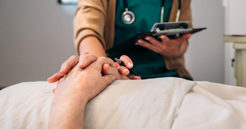 A nurse touches the hands of an elderly male patient laying in a hospital bed.