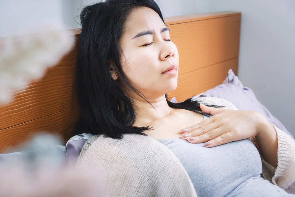 Una mujer asiatica acostada en una cama con dolor y la mano en el pecho.