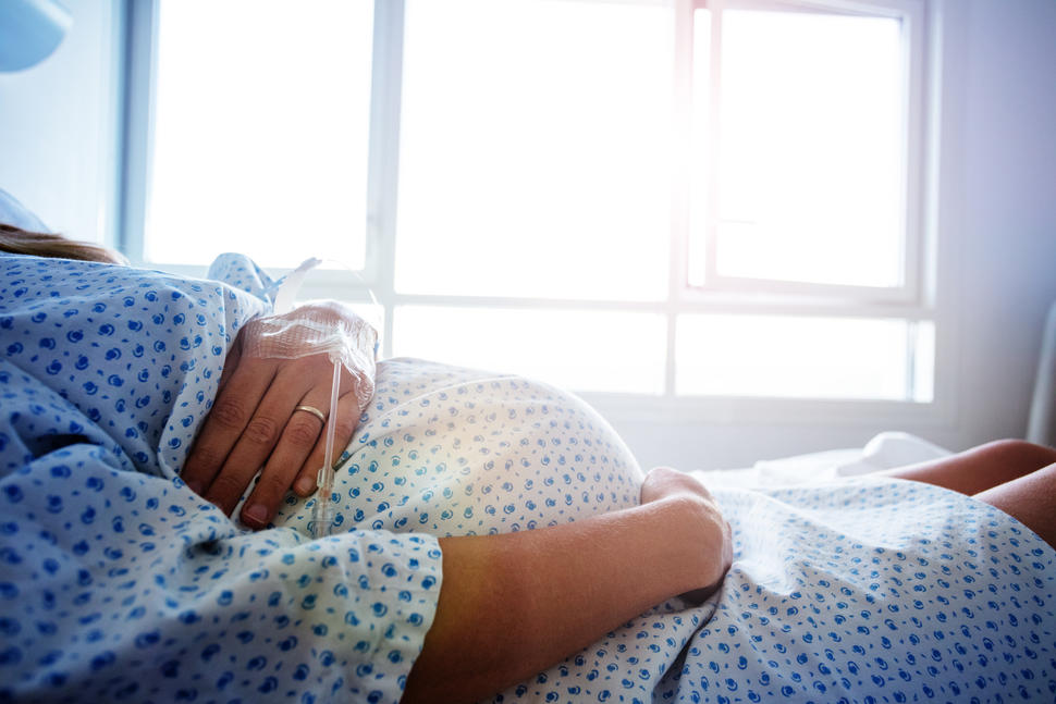 Foto de medio cuerpo de una mujer embarazada puesta una bata de hospital con una mano sobre su barriga, en la misma mano tiene un IV.