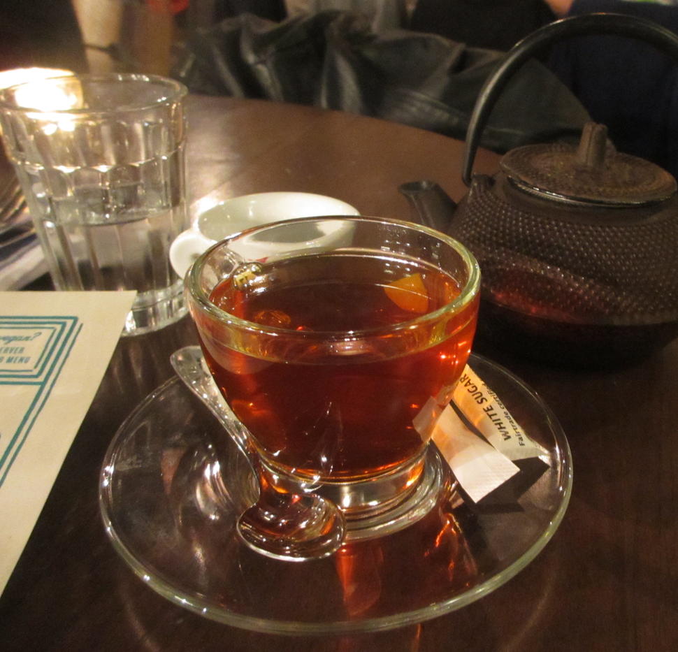 A cup of Black tea with a spoon and packet of sugar sitting on a saucer. A teapot sits behind the cup.  