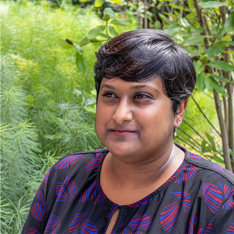 Deepa Prasad stands in front of a green, wooded background while looking at something off camera to her right. She is a medium-skinned woman with short, dark hair and is wearing a dark blue top with a light purple brushstroke print. 