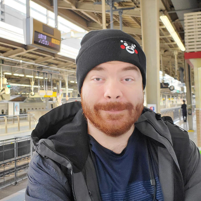 Headshot of a light-skinned man, Justin, in a black hat, black and grey jacket, and blue shirt