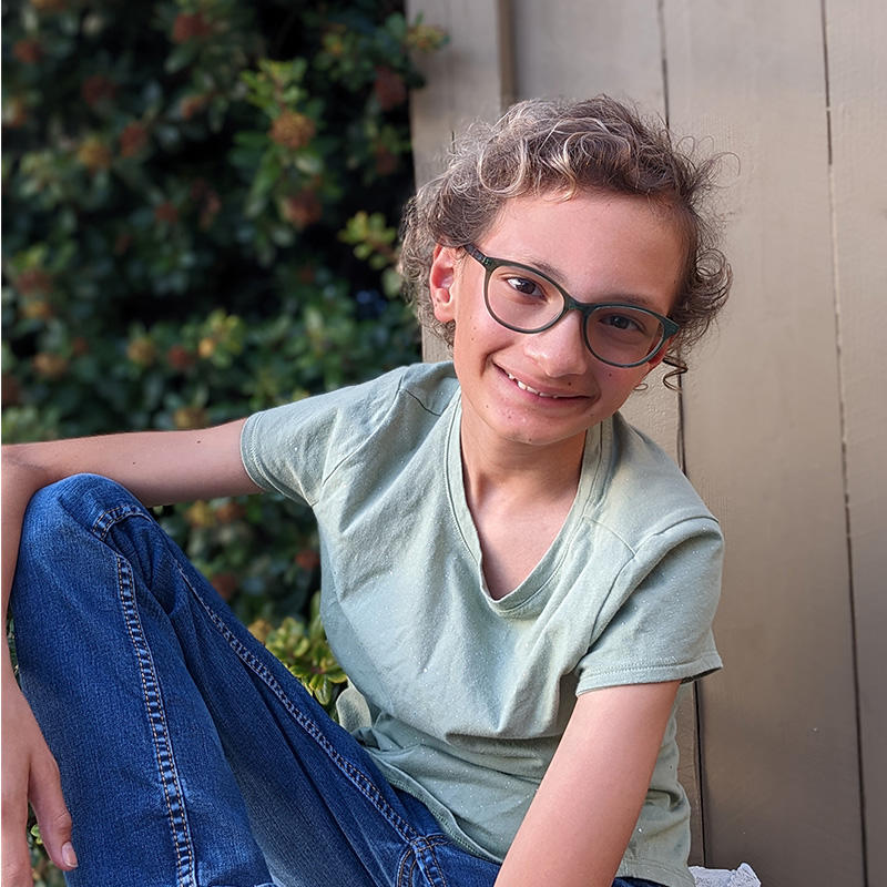 Micah is a young, light-skinned, non-binary person with short curly hair and glasses. They are wearing a mint green t-shirt and jeans and are smiling at the camera. 