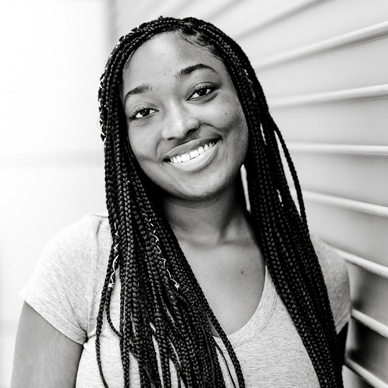 Black and white photo of young, Black woman with braids, standing against a building.