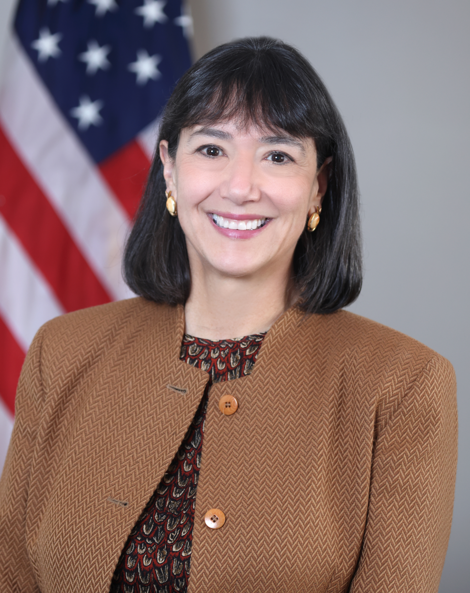 Woman (NCI Director Dr. Monica Bertagnolli) with dark hair wears a brown jacket over a patterned top and smiles at the camera.