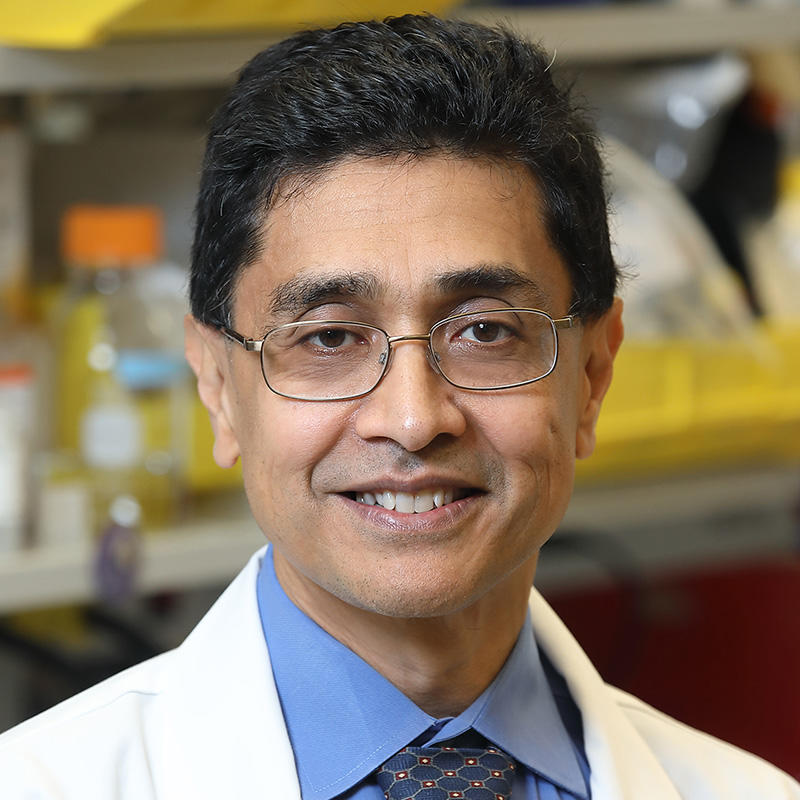 A man with medium brown skin tone and dark hair, wearing glasses, a blue shirt, a navy-blue tie, and a lab coat, is standing in a lab smiling at the camera.