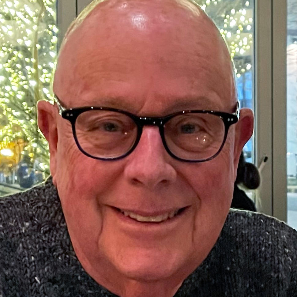 White man with glasses and grey knit sweater smiling at camera while sitting in front of glass doors. Outside are trees covered in white lights.