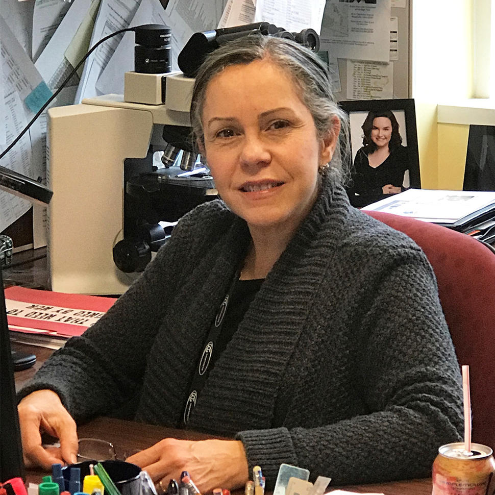 Puerto Rican woman (Nilsa Ramirez) sits at a desk with a microscope, framed photo, and papers tacked to a bulletin board behind her.