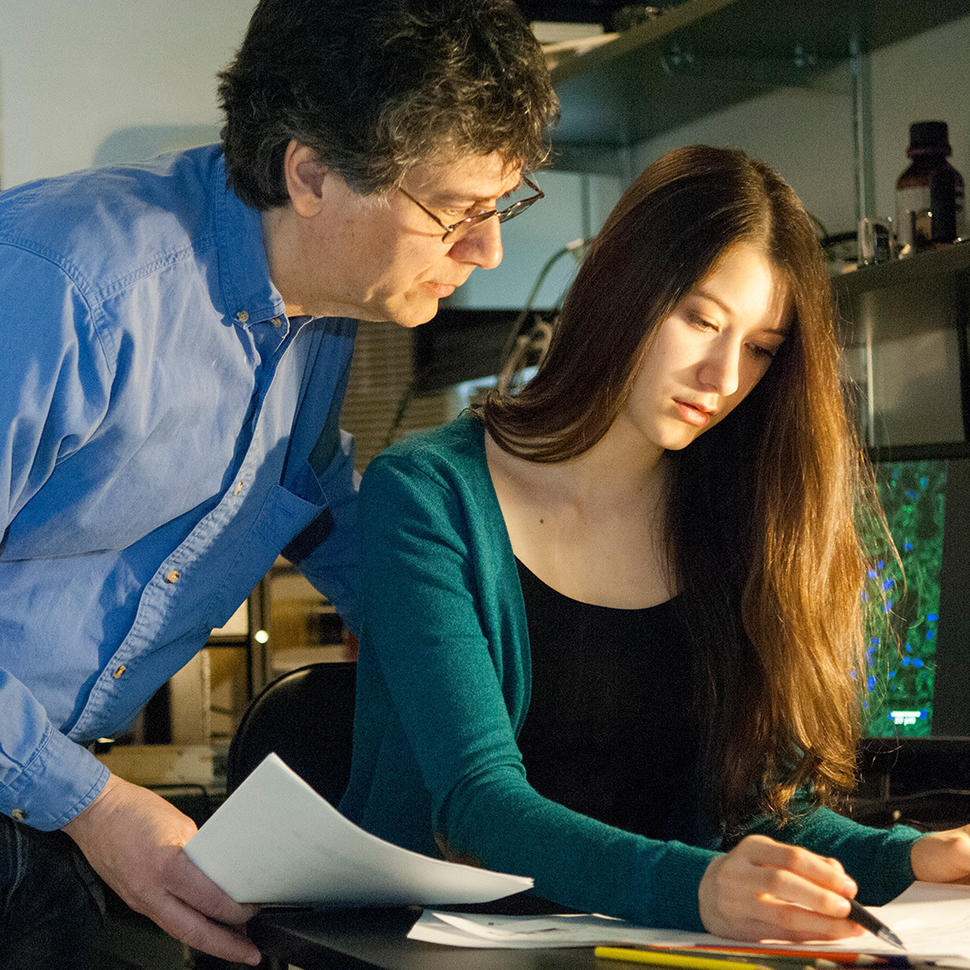 A young woman sits looking and pointing a pen at a paper on a desk in a lab office. Her father leans over her shoulder looking at the same paper. 