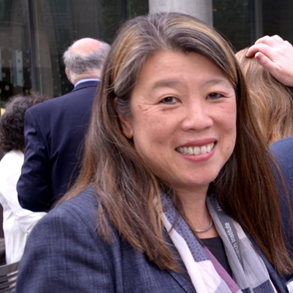 An Asian woman with light skin tone wearing a blue blazer (Dr. Mignon Lee-Cheun Loh) smiles at the camera at an outdoor conference event. 