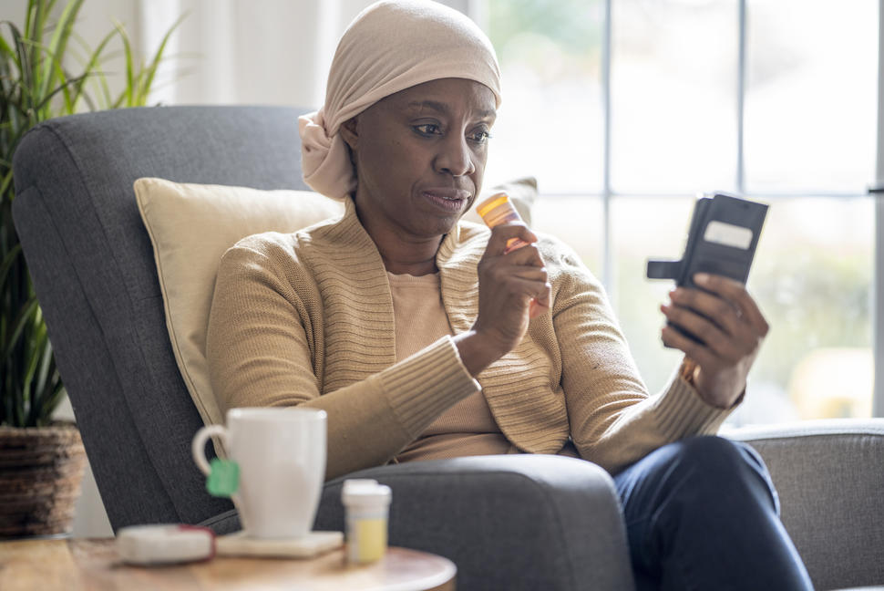 Woman wearing a head scarf holding a bottle of medication in one hand and looking at her phone in the other hand.