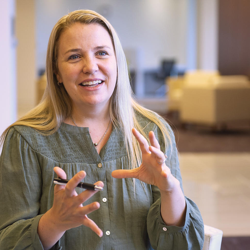 A woman (Dr. Emily Tonorezos) with long blond hair wears a muted green button-up blouse and sits in a chair, smiling and talking with her hands.