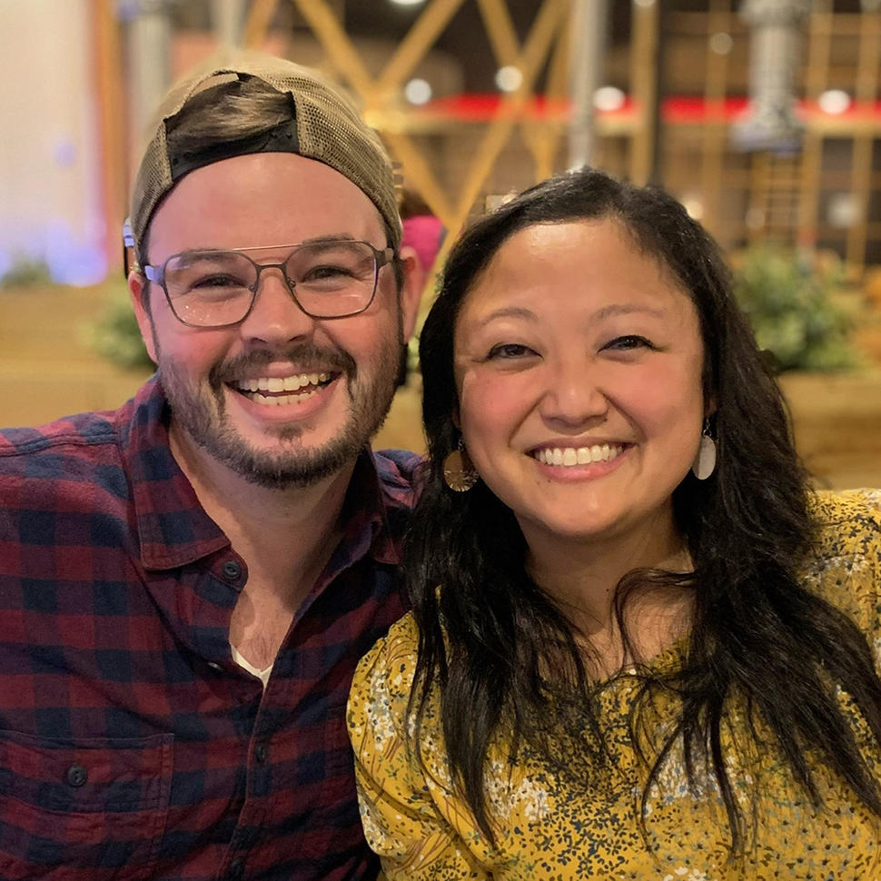 Woman (Sarah) with long black hair, earrings, and peach-colored blouse with blue and white flowers sits next to her husband. Both smile at the camera.