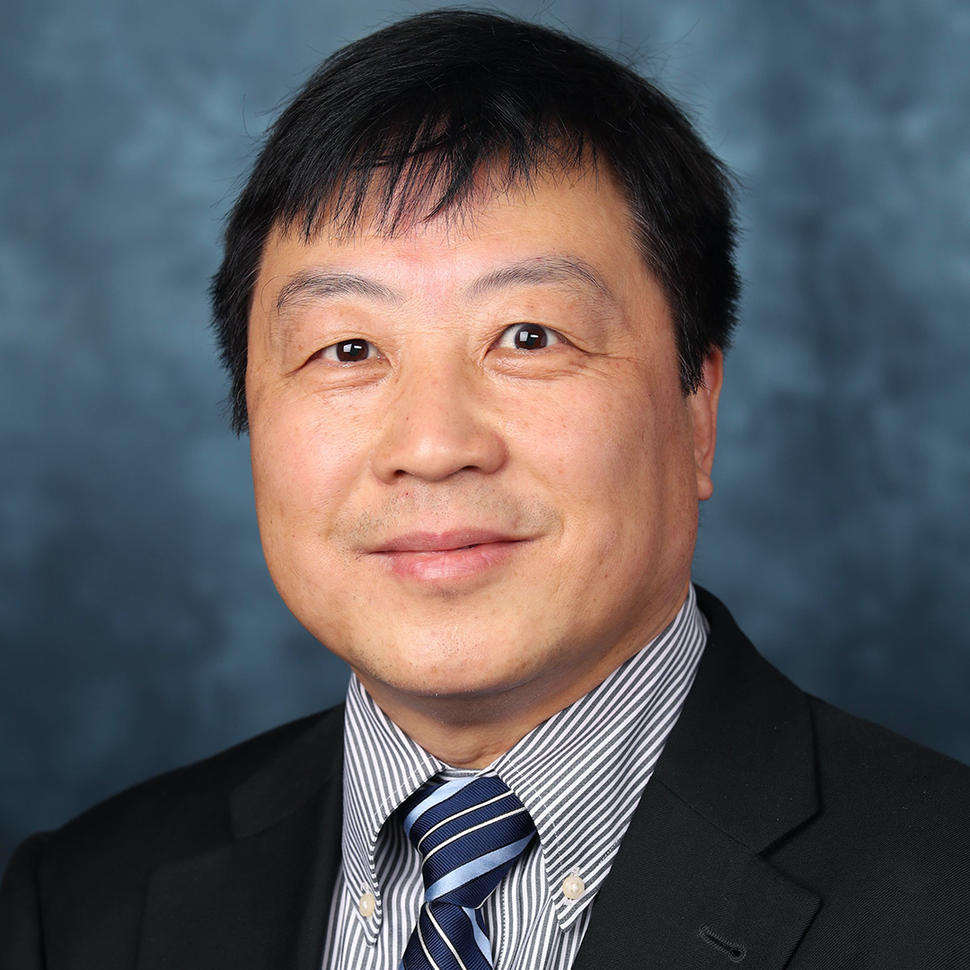 A man (Dr. Xiao-Nan Li) with dark hair and brown eyes wears a striped collared shirt, tie, and dark jacket and smiles at the camera.