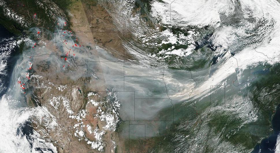 A satellite image of the United States with visible smoke plumes extending across several states. Numerous red dots indicate active wildfire locations, predominantly in the Pacific Northwest and Northern California. The smoke appears to be drifting eastward, covering parts of the Midwest. The image captures a mix of cloud cover and clear areas, with some mountainous regions visible.