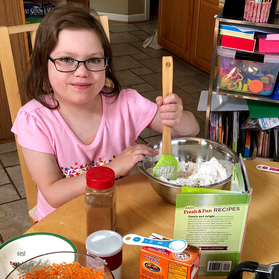 A girl, Abby, wearing glasses and a pink T-shirt, stirs ingredients in a metal bowl with a rubber spatula and smiles at the camera.