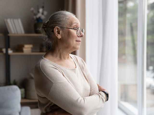 Woman standing indoors by a window, smiling and gazing outside.