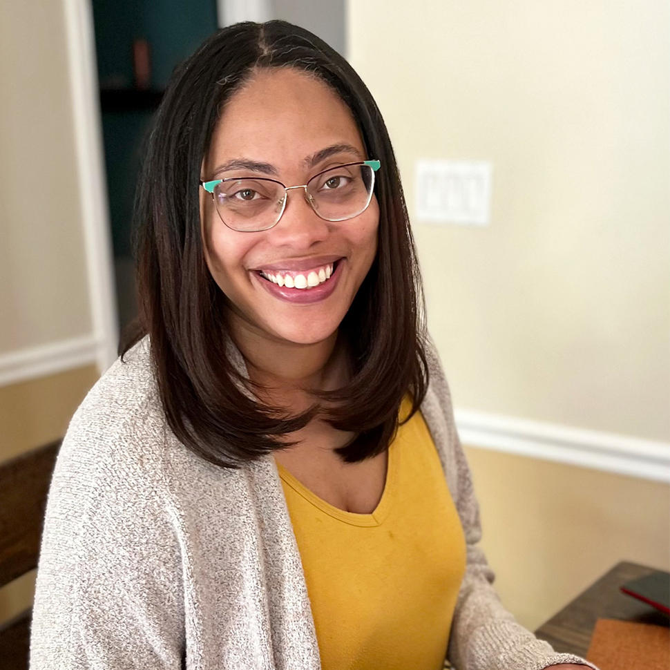 A woman, Dr. Robin Lockridge, with glasses and straight brown hair wearing a yellow shirt and gray cardigan sits at her laptop smiling at the camera.