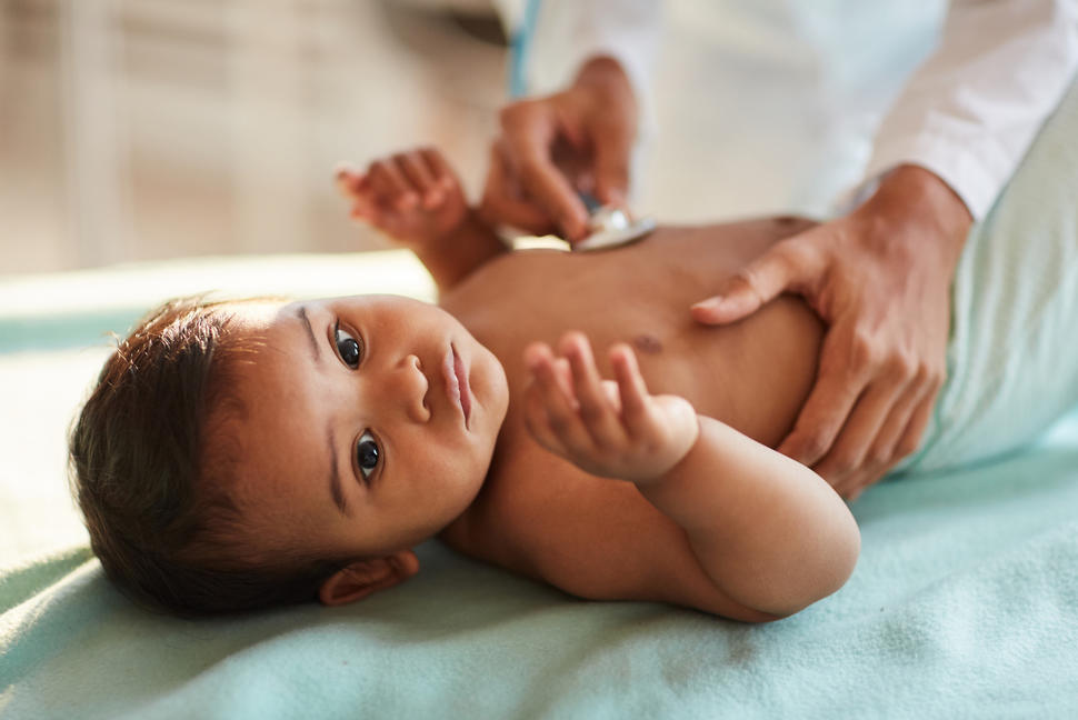 Photo of an infant lying on an exam table with an adult holding a stethoscope to the baby's chest.