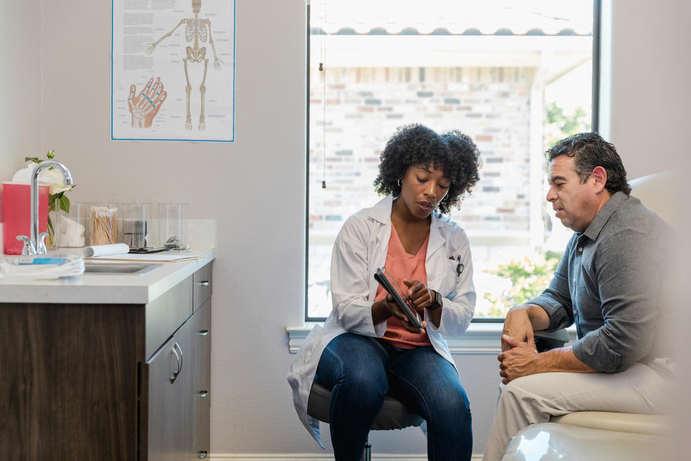 A female doctor points to something on tablet as a male patient listens.