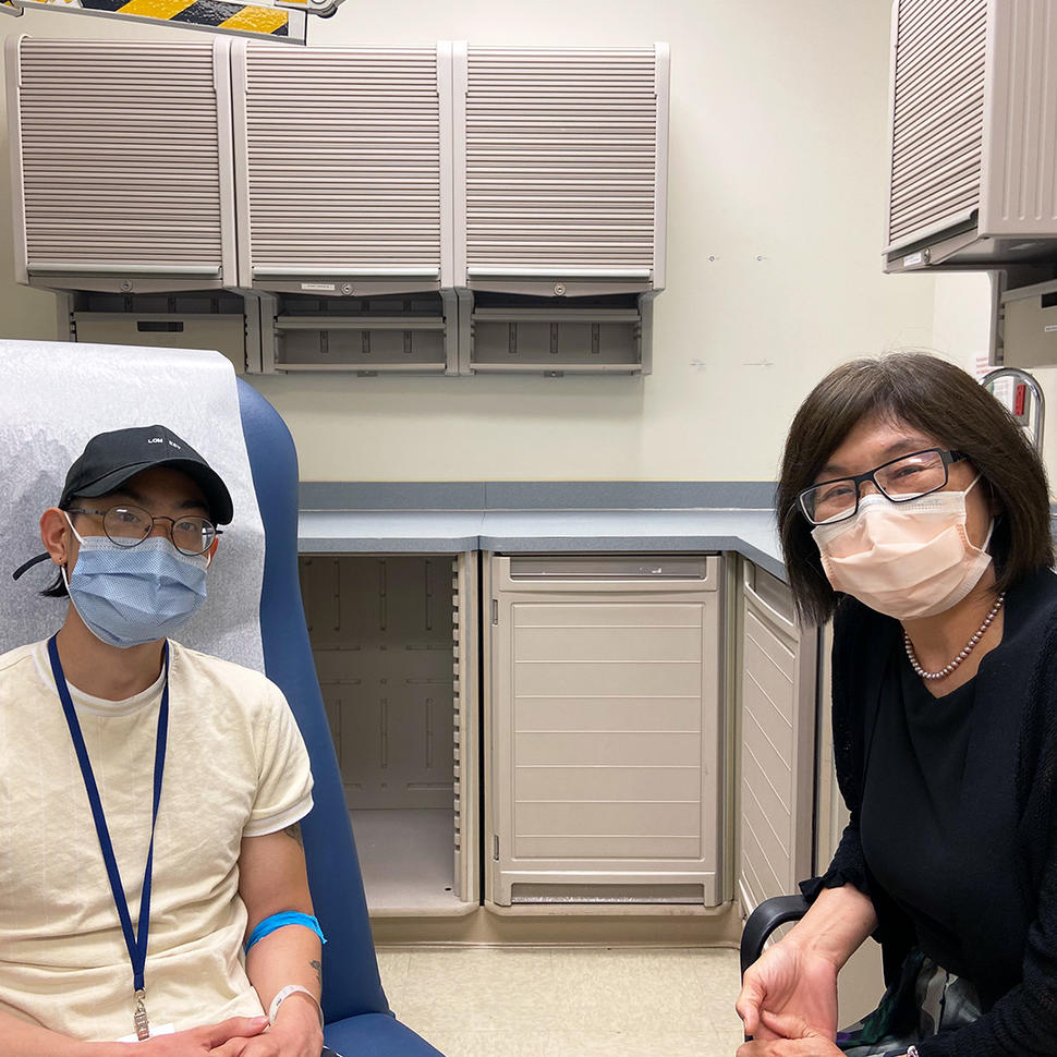 A doctor, Dr. Alice Chen, with short, dark hair wearing glasses and a surgical mask sits next to a patient wearing a hat, glasses, surgical mask, and yellow t-shirt in an exam room.