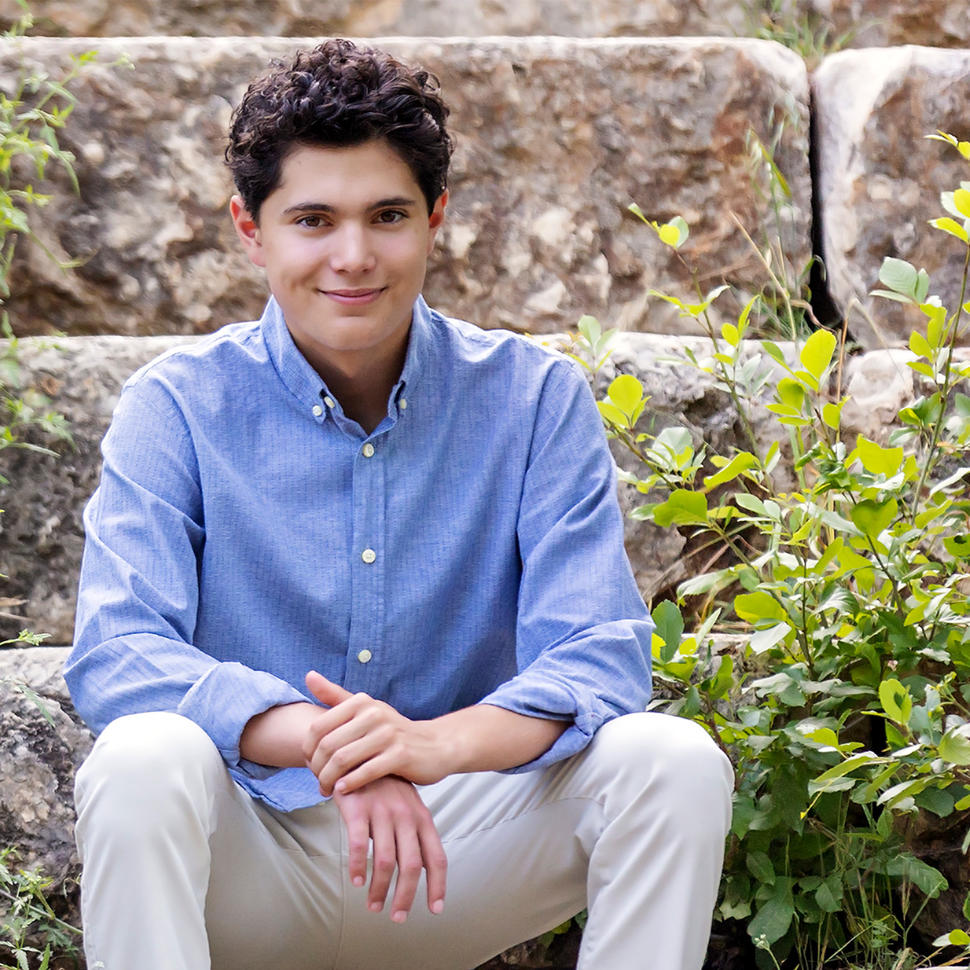 A young man, Callan, with brown hair and eyes, sits on a stone step wearing a blue button-up shirt and dress pants and smiles at the camera.