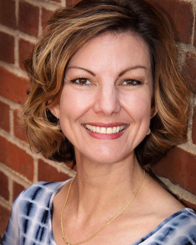 Woman with short, brown hair wearing a blue and white top smiling at the camera while leaning against a brick wall.