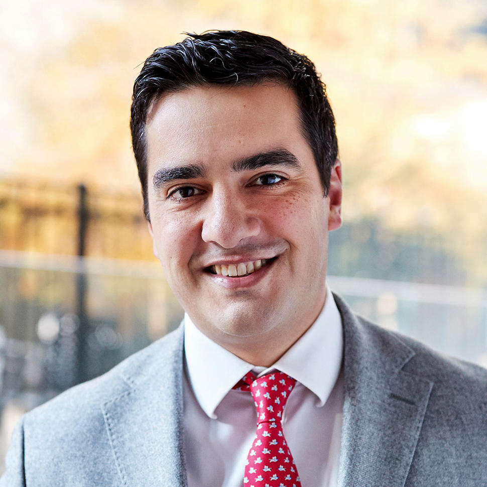 A man, Dr. Michael Ortiz, with brown hair and brown eyes and wearing a grey suit with a red tie, smiles at the camera.