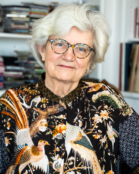 Female woman with black multi color blouse with bookshelf behind her.